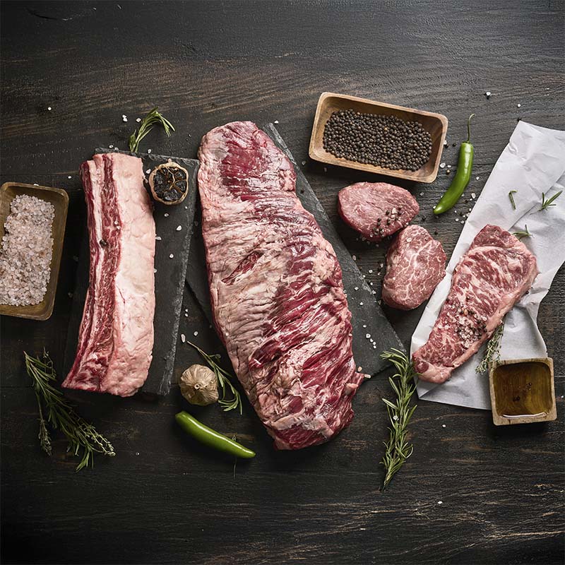 A collection of steaks featured in the premium mix bundle on a wood table, surrounded by fresh herbs, peppers, and spices.