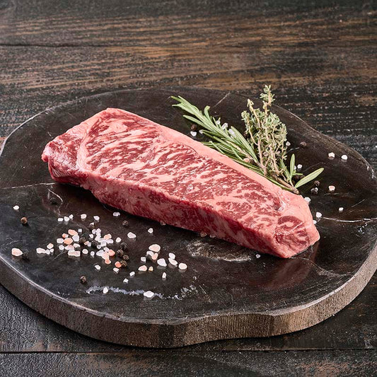 A beautifully marbled NY Strip Steak from Yolo 8 Meats on a dark wood cutting board, surrounded by rock salt and fresh herbs.