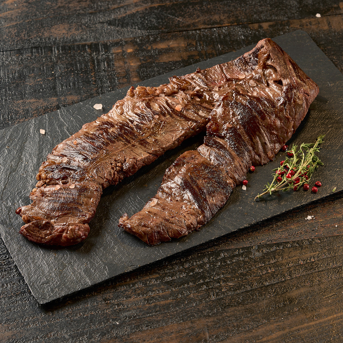 A grilled skirt steak on a dark rectangular serving board, with a small herb garnish, photographed on a rustic dark wooden table.
