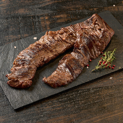 A grilled skirt steak on a dark rectangular serving board, with a small herb garnish, photographed on a rustic dark wooden table.