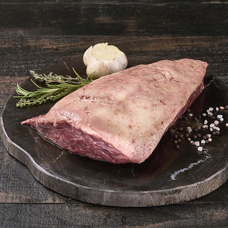 A picanha roast with a fat cap, sitting on a black wood cutting board surrounded by fresh garlic and herbs.