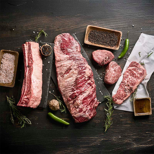 A collection of steaks featured in the premium mix bundle on a wood table, surrounded by fresh herbs, peppers, and spices.
