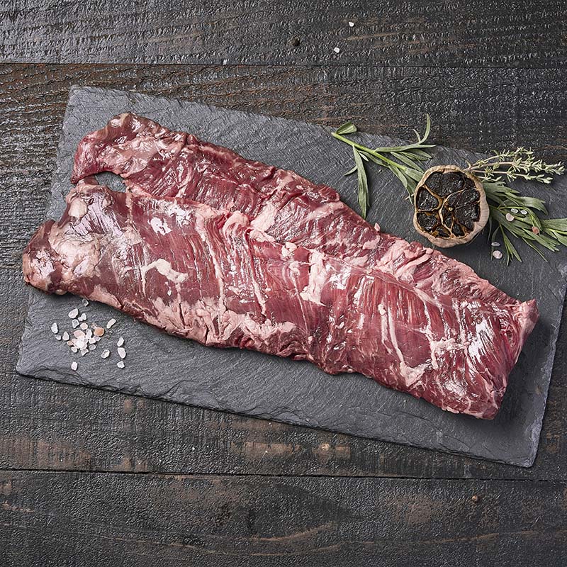 A long strip of skirt steak, doubled over on itself, sitting on a black stone platter surrounded by fresh herbs, rock salt, and roasted black garlic