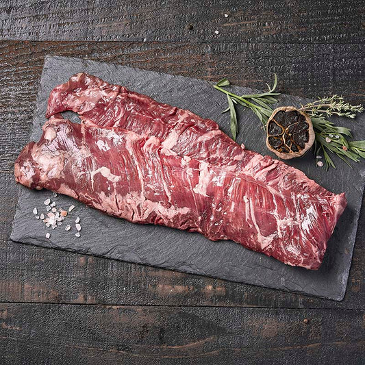 A long strip of skirt steak, doubled over on itself, sitting on a black stone platter surrounded by fresh herbs, rock salt, and roasted black garlic