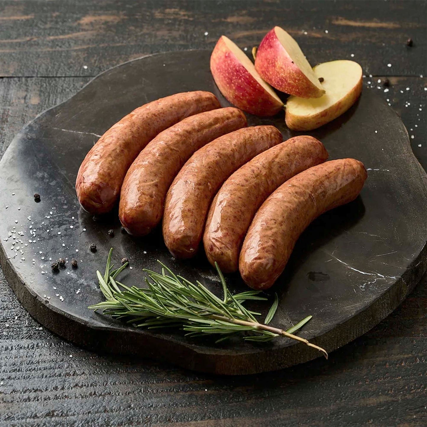 Five sausages on a dark stone plate with apple slices and rosemary on a wooden background