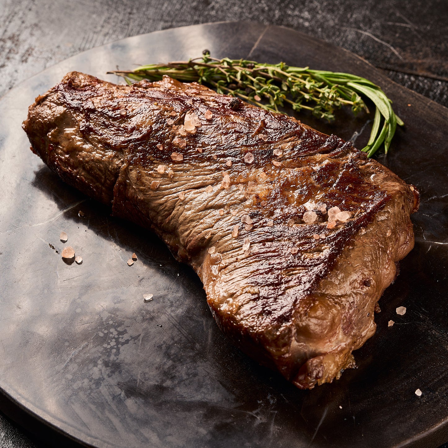 A seared tri tip steak with visible salt crystals, served on a dark plate with a sprig of rosemary against a dark textured background.