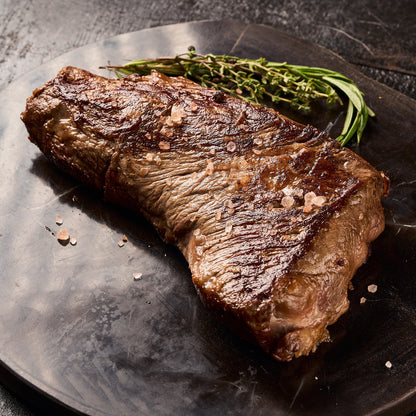 A seared tri tip steak with visible salt crystals, served on a dark plate with a sprig of rosemary against a dark textured background.
