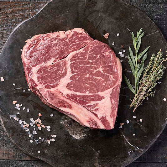 A beautifully marbled bone-in ribeye steak from Yolo 8 Meats on a dark wood cutting board, surrounded by rock salt and fresh herbs.