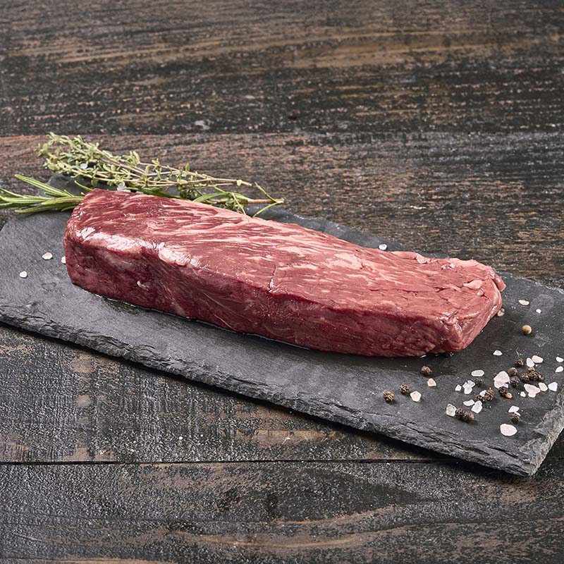 A stout Denver Steak with ribbons of fat marbling, sitting on a black stone platter surrounded by rock salt, peppercorns, and fresh herbs.
