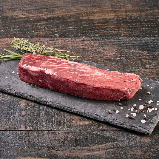 A stout Denver Steak with ribbons of fat marbling, sitting on a black stone platter surrounded by rock salt, peppercorns, and fresh herbs.