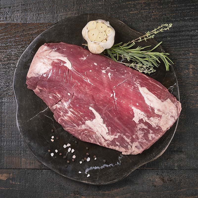 A slab of flank steak sitting on a dark wood cutting board, surrounded by fresh garlic and herbs.
