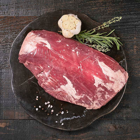 A slab of flank steak sitting on a dark wood cutting board, surrounded by fresh garlic and herbs.
