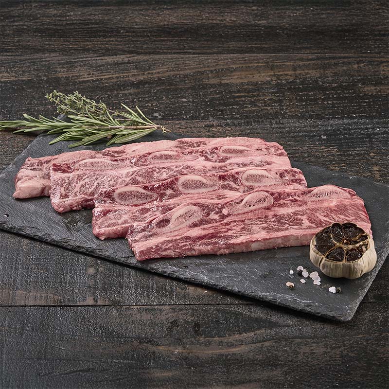 A set of 4 flat flanken ribs with exquisite marbling, on a black stone surface with black garlic, rock salt, and fresh herbs surrounding.