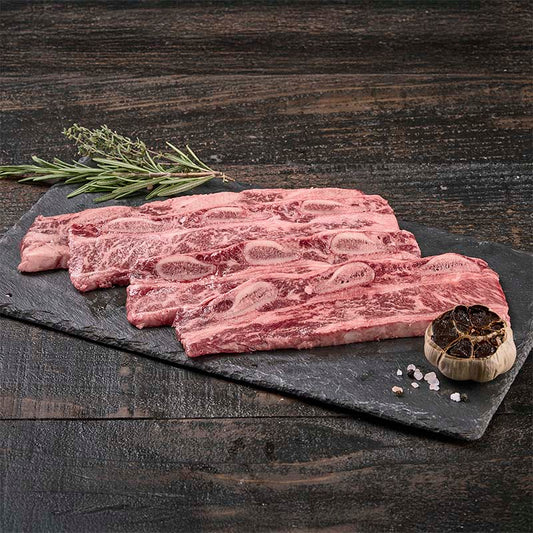 A set of 4 flat flanken ribs with exquisite marbling, on a black stone surface with black garlic, rock salt, and fresh herbs surrounding.