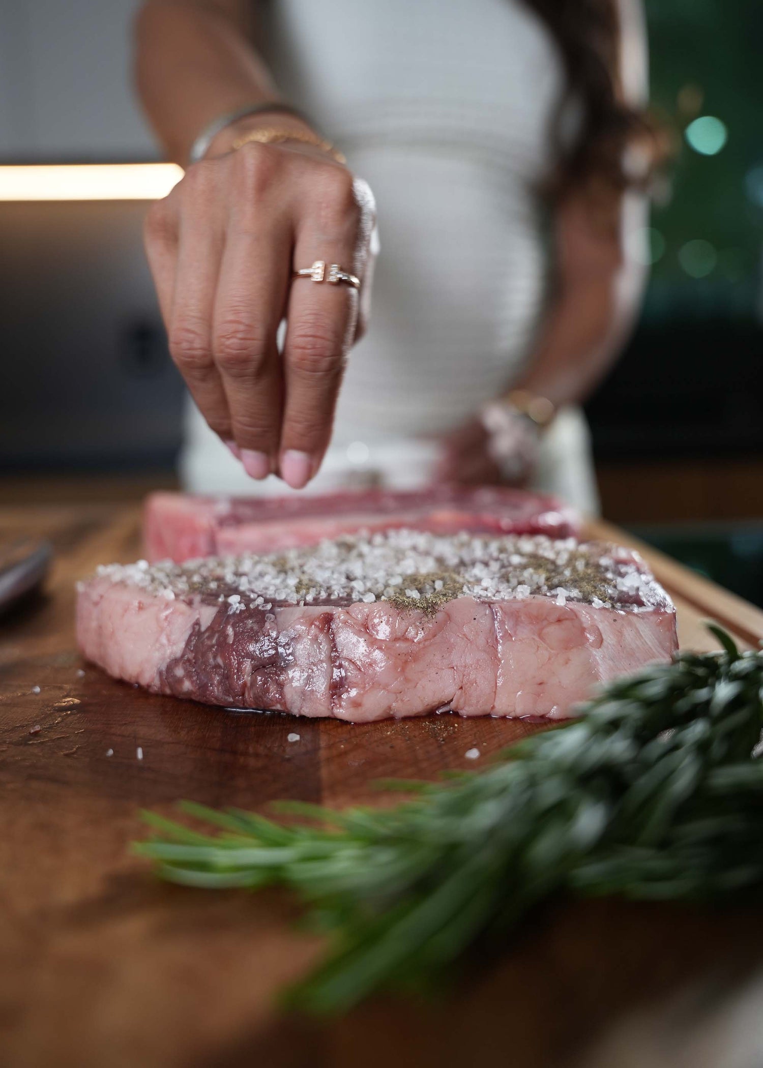 Woman seasoning a piece of meat on a wooden cutting board with herbs.