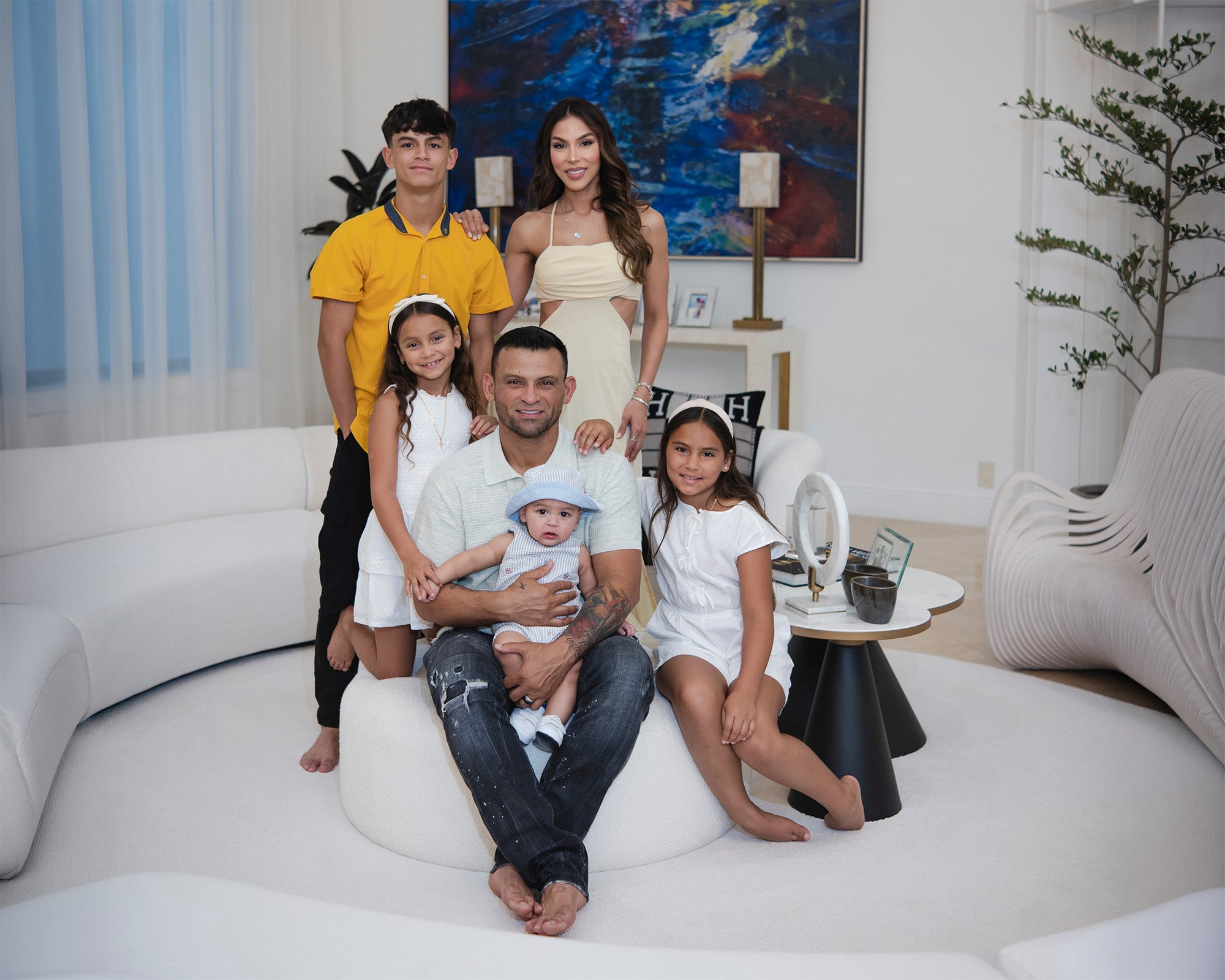 A family of six posing together in a modern, minimalist living room with white curved sofas and a large abstract painting on the wall. The father is sitting on a white circular ottoman in the center, holding a baby, while the mother stands behind them in a cream dress.