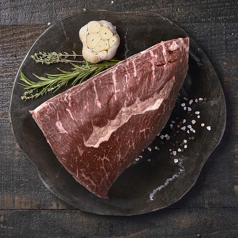 A picanha roast, sitting on a black wood cutting board surrounded by fresh garlic and herbs.