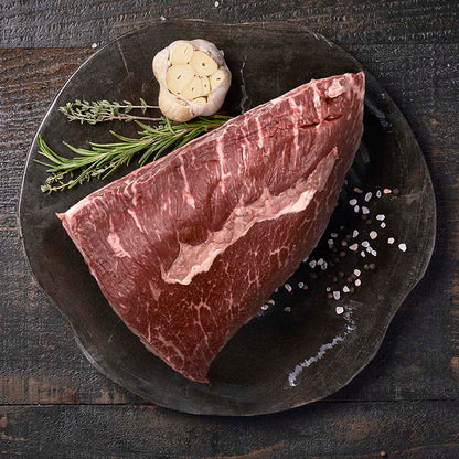 A picanha roast, sitting on a black wood cutting board surrounded by fresh garlic and herbs.