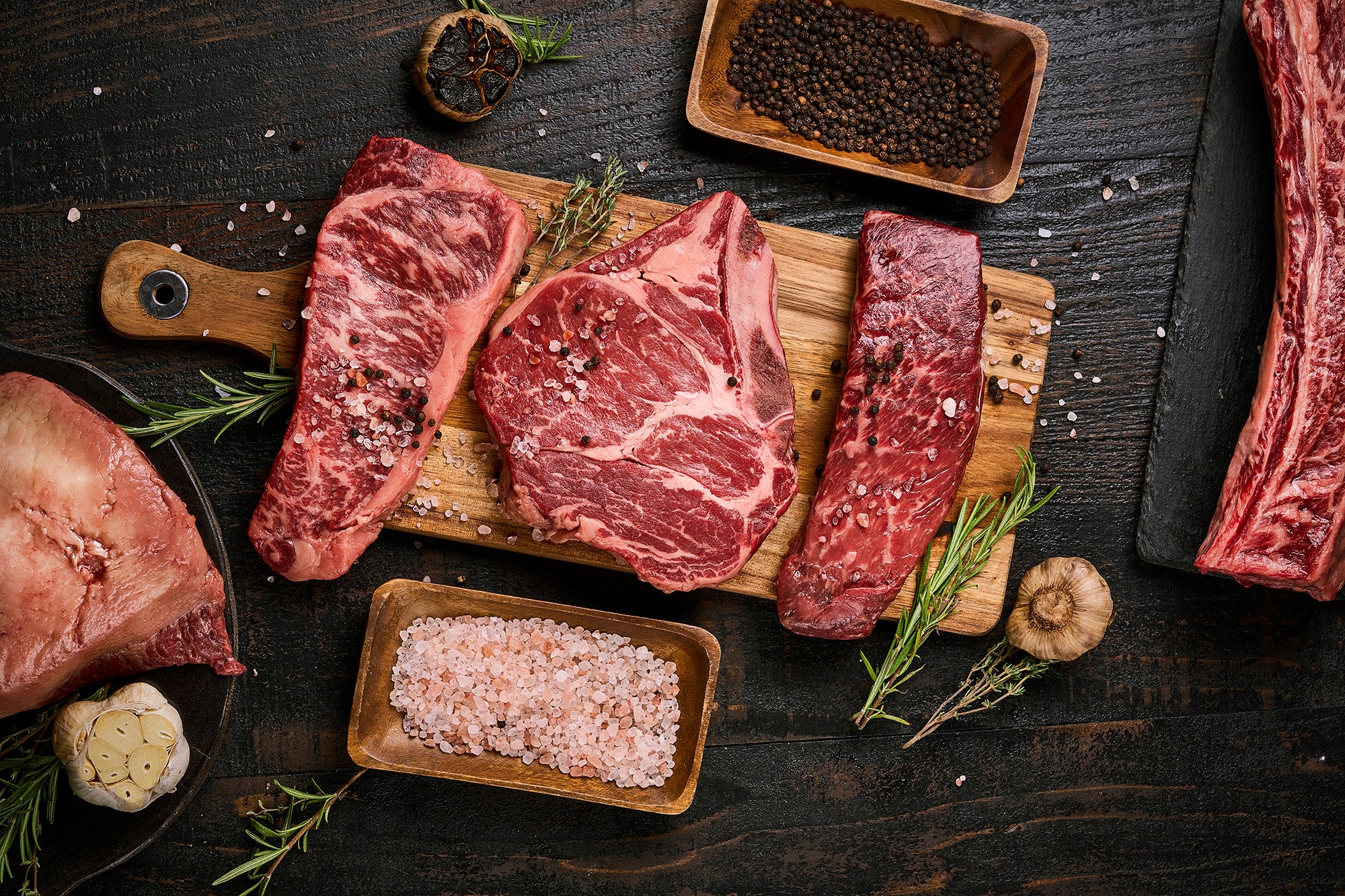 Raw steaks on a wooden cutting board with salt, pepper, and rosemary on a dark surface.