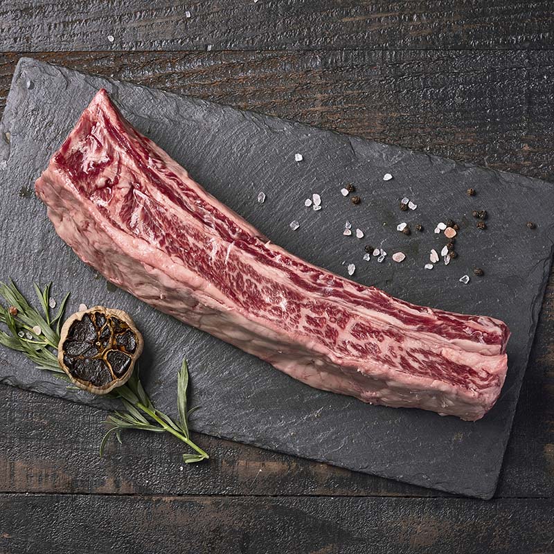 Exquisitely marbled beef short ribs from Yolo 8 Meats, sitting on a black stone platter surrounded by roasted black garlic, fresh herbs, and rock salt.