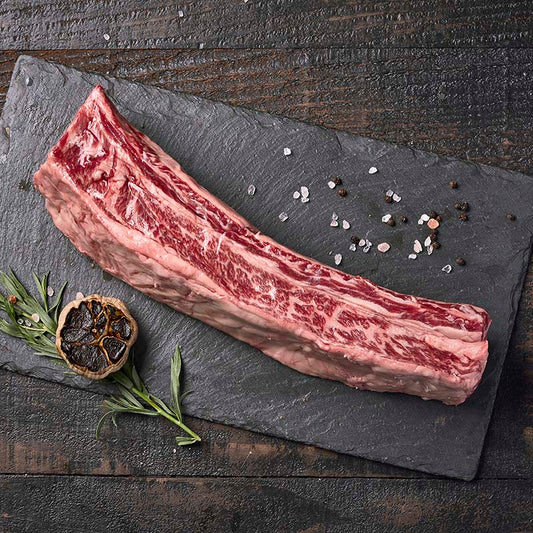 Exquisitely marbled beef short ribs from Yolo 8 Meats, sitting on a black stone platter surrounded by roasted black garlic, fresh herbs, and rock salt.